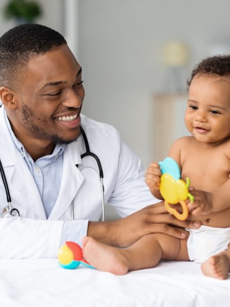 Portrait Of Smiling Black Doctor Making Medical Check Up For Little Baby Patient In Clinic, Handsome Young African American Pediatrist Looking At Adorable Infant Child In Diaper, Closeup Shot